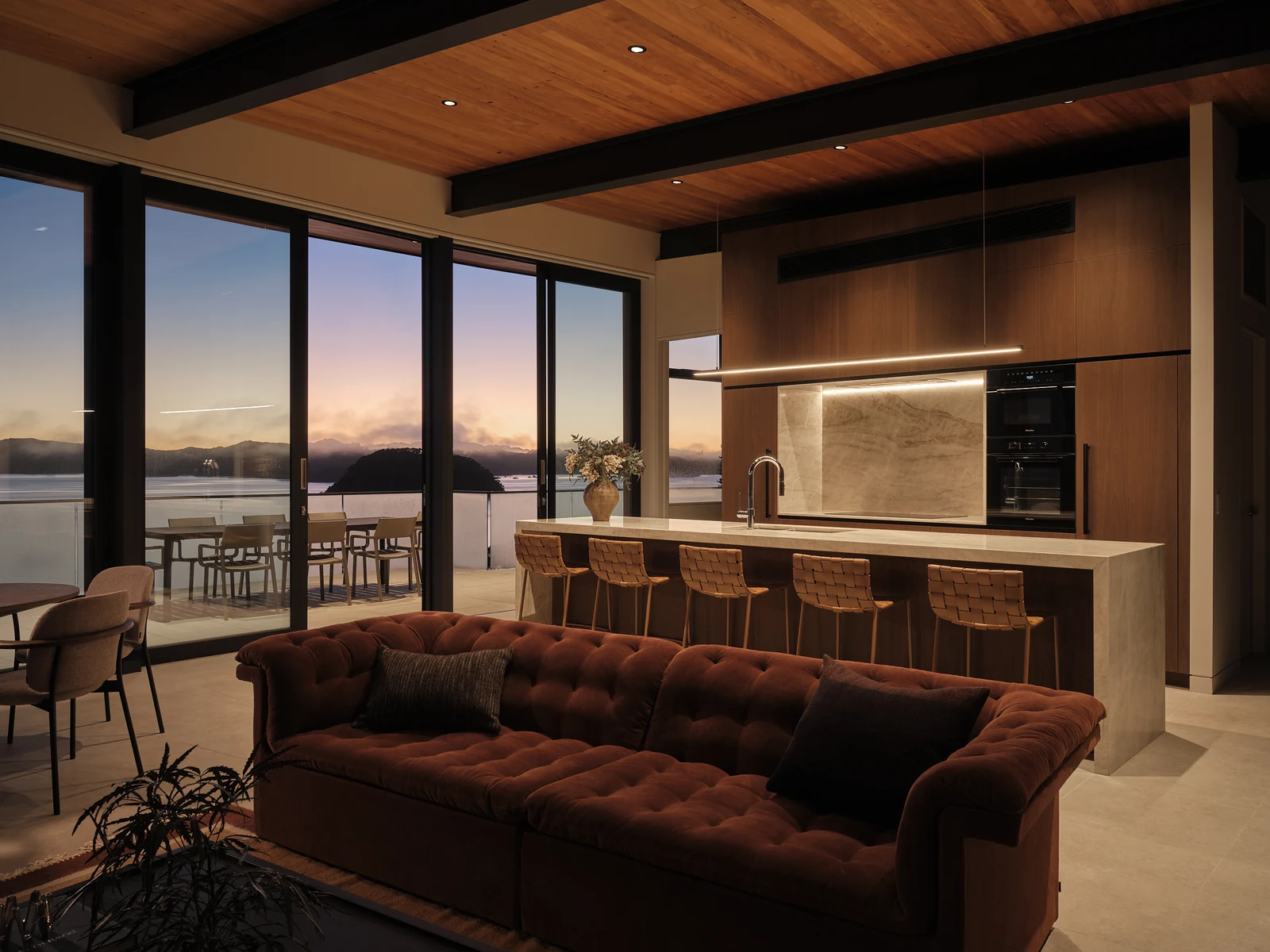 A wide view of the kitchen and living room before dawn, with warm cabinetry lighting and blue sky over the bay