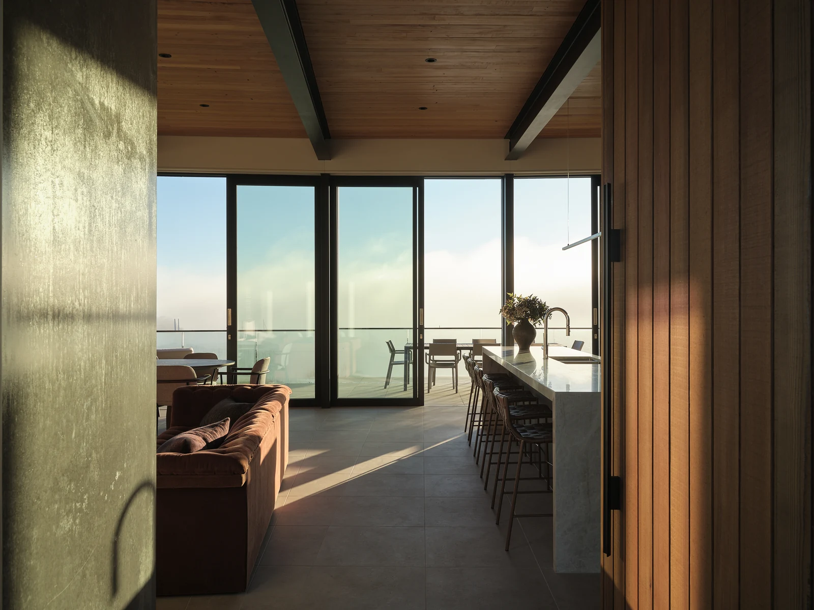 Morning light along the School Road kitchen island towards the terrace and bay, framed by timber and a textured wall