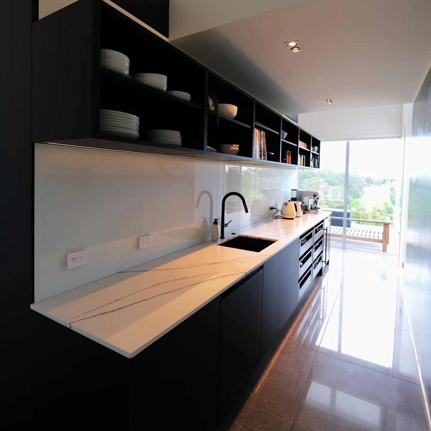 Black galley kitchen with white stone worktop, open shelving and a long garden outlook