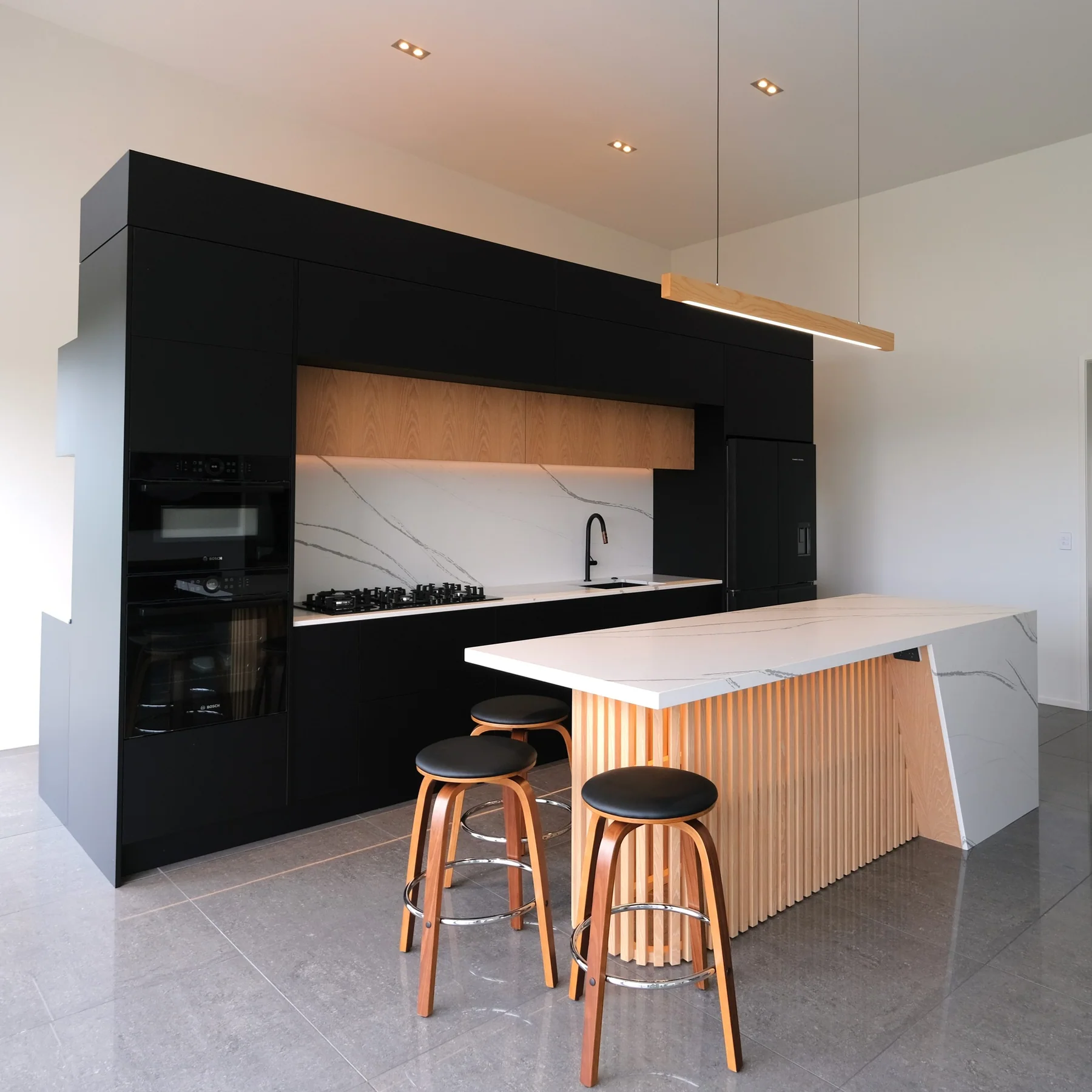 Black kitchen wall with a pale stone and timber-slat island, pendant lighting and timber stools