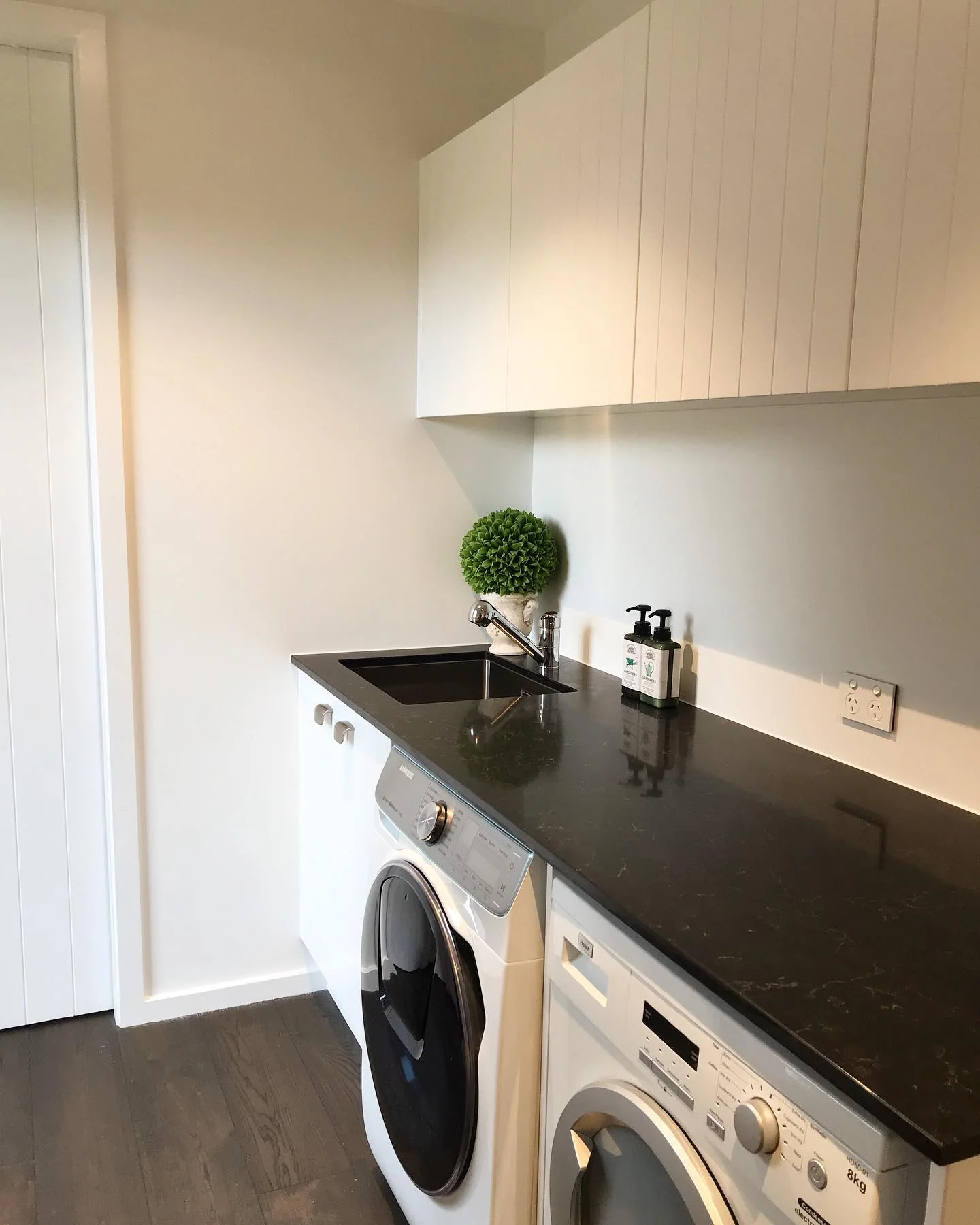 Compact white laundry with black stone benchtop, integrated sink, overhead storage, washer and dryer