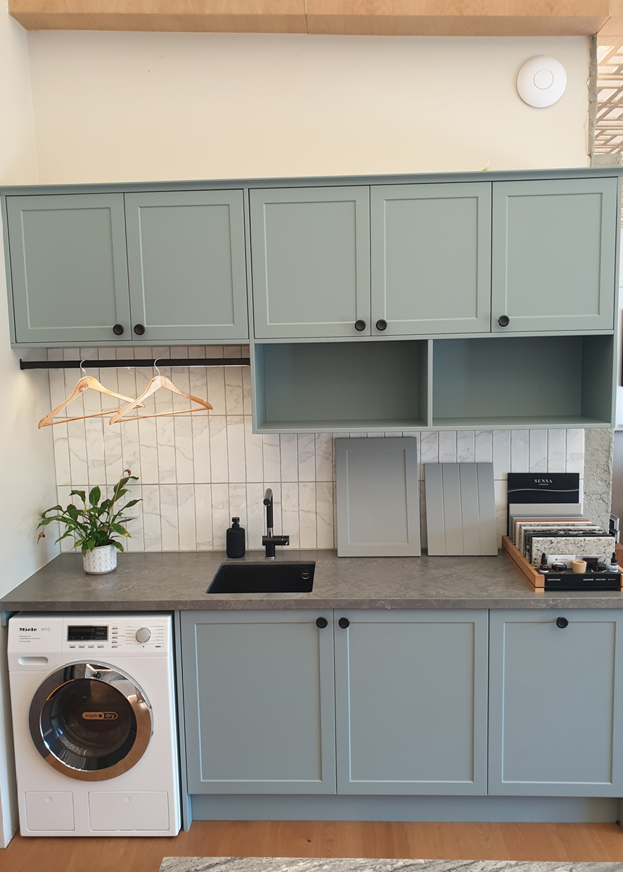 Sage green Shaker laundry cabinetry with sink, hanging rail, stone benchtop and integrated washer by North Interiors