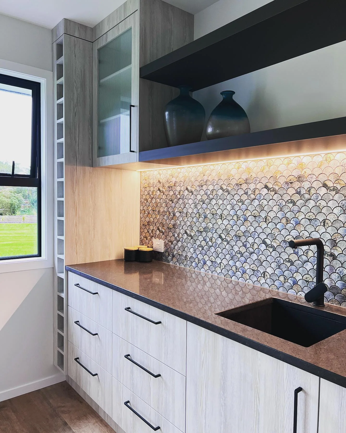 Light timber scullery cabinetry with black worktop, open shelving and a shell-pattern tiled splashback